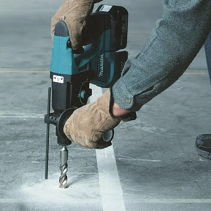 Makita LXT 18V Akku-Kombihammer DHR243RTJ - 18 V, 2 Akkus, 5 Ah, 2 J, Ohne Staubabsaugung - Ein Makita Akku-Bohrhammer wird von einer Person mit Handschuhen bedient, um ein Loch in einen Betonboden zu bohren.
