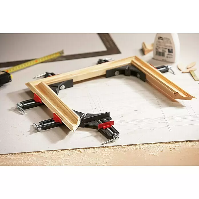 Bessey Winkelspanner WS1 - Spannweite: 73 mm - Schwarze und rote Eckzwingen halten Gehrungshölzer eines Bilderrahmens auf einer Werkbank zusammen.