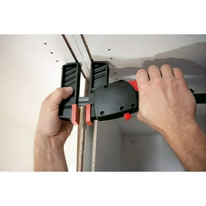 Bessey Einhandzwinge DuoKlamp DUO 30-8 - Spannweite: 300 mm, Spannkraft: 1.200 N, Ausladung: 85 mm - Einhandzwinge im Einsatz, um zwei weiße Bretter zu fixieren. Das Werkzeug ist schwarz mit roten Akzenten und wird von zwei Händen bedient.