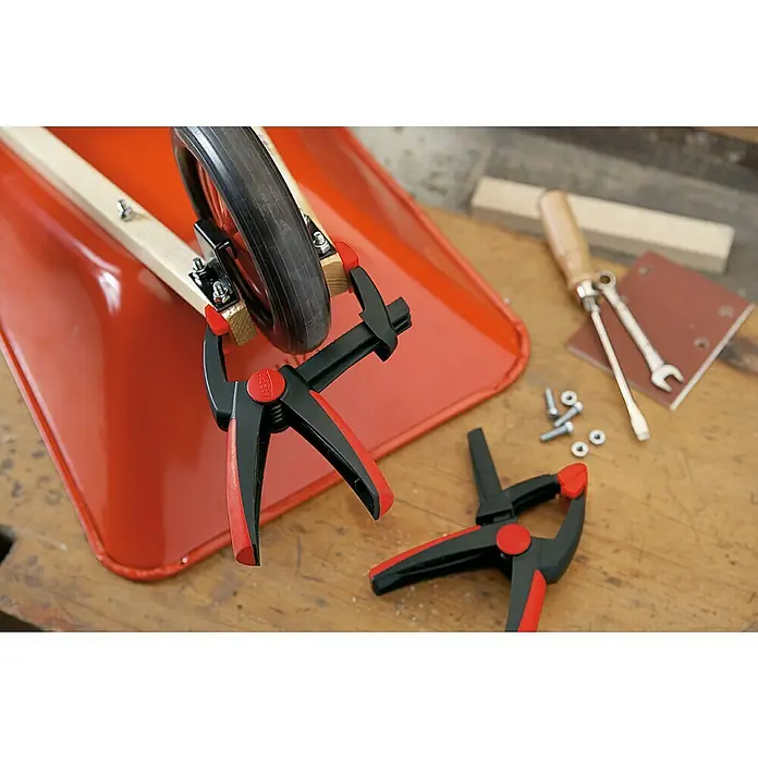 Bessey Federzwinge Clippix Vario XV 5-100 - Spannweite: 100 mm, Spannkraft: 120 N, Ausladung: 50 mm - Detailaufnahme einer Werkbank mit rot-schwarzen BESSEY-Knebelhebelzwingen, die ein Rad an einer Holzkonstruktion befestigen. Werkzeuge liegen bereit.