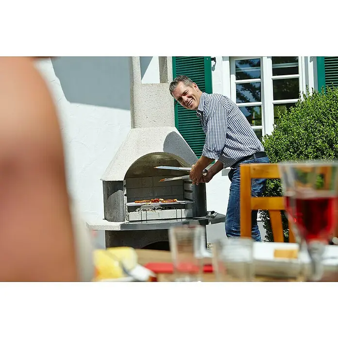 Buschbeck Grillkamin Stockholm - Hauptgrillfläche: 54 x 34 cm - Ein Mann grillt im Freien, wobei der Fokus auf dem Grill und den darauf zubereiteten Speisen liegt.