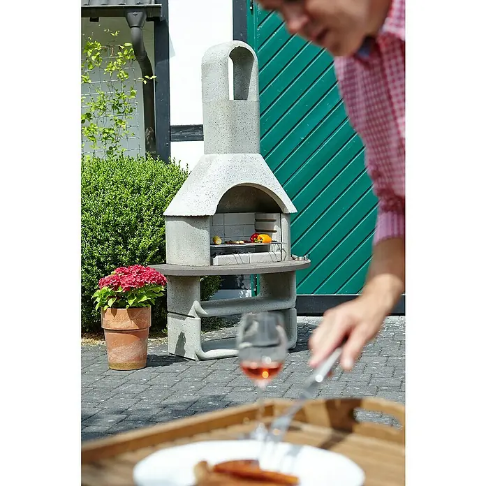 Buschbeck Grillkamin Capri - Hauptgrillfläche: 54 x 34 cm, Feuerfester Leichtbeton - Ein gemauerter Gartengrill mit Speisen auf dem Grillrost neben einem gedeckten Tisch.