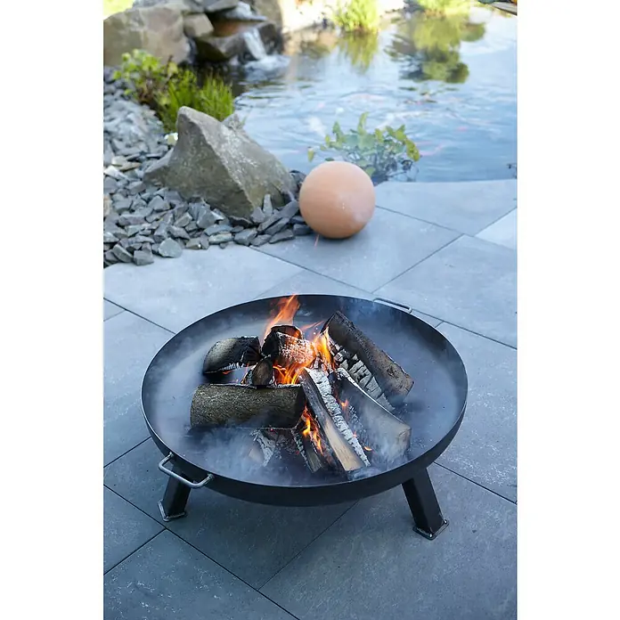 Buschbeck Feuerschale - Durchmesser: 55 cm, Eisen - Feuerstelle im Freien mit brennendem Holz und einem Teich im Hintergrund. Ideal für gemütliche Abende im Garten.