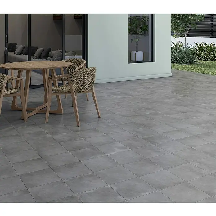 Pavimento porcelánico Madox - 33,3 x 33,3 cm, Antracita, Mate - La imagen muestra un elegante espacio exterior con baldosas grises, destacando una elegante zona de comedor con mesa de madera y sillas de mimbre.