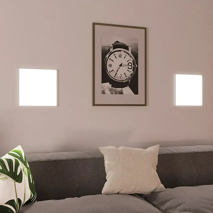 Ledvance SUN@Home LED-Panel Planon Frameless - 20 W, L x B x H: 30 x 30 x 6,1 cm, Weiß, Kaltweiß - Wandleuchten neben einem gerahmten Bild über einer Couch mit Kissen. Die quadratischen Lichter erhellen den Raum und betonen das Dekor.