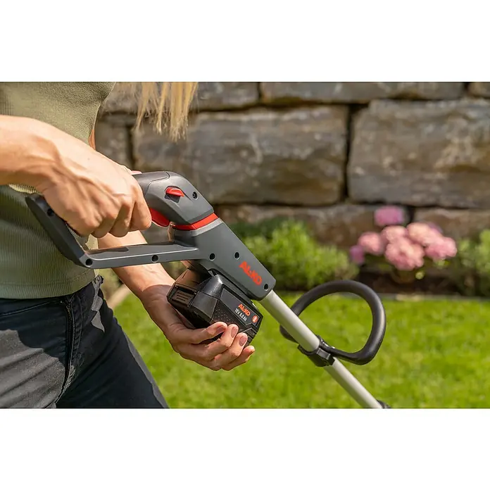 AL-KO Bosch Home & Garden Compatible Akku-Rasenlüfter AR 1835 SET - Anzahl Akkus: 1 Akku, 18 V, Arbeitsbreite: 35 cm - Nahaufnahme eines AL-KO Akku-Rasentrimmers mit eingesetztem Akku vor einem Garten mit Blumen.