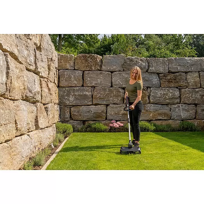 AL-KO Bosch Home & Garden Compatible Akku-Rasenlüfter AR 1835 SET - Anzahl Akkus: 1 Akku, 18 V, Arbeitsbreite: 35 cm - Ein schwarz-grauer Rasenkehrer wird von einer Frau auf einem grünen Rasen an einer Steinmauer benutzt.