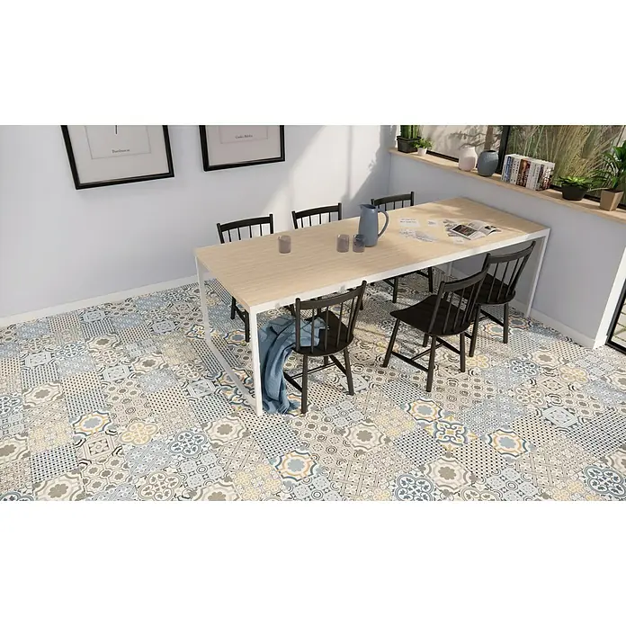 Pavimento porcelánico hidráulico Windsor - 45 x 45 cm, Azul/Gris/Crema, Mate - Comedor con mesa de madera clara rodeada de sillas negras, sobre un suelo de baldosas patchwork. Elementos decorativos expuestos en el alféizar.
