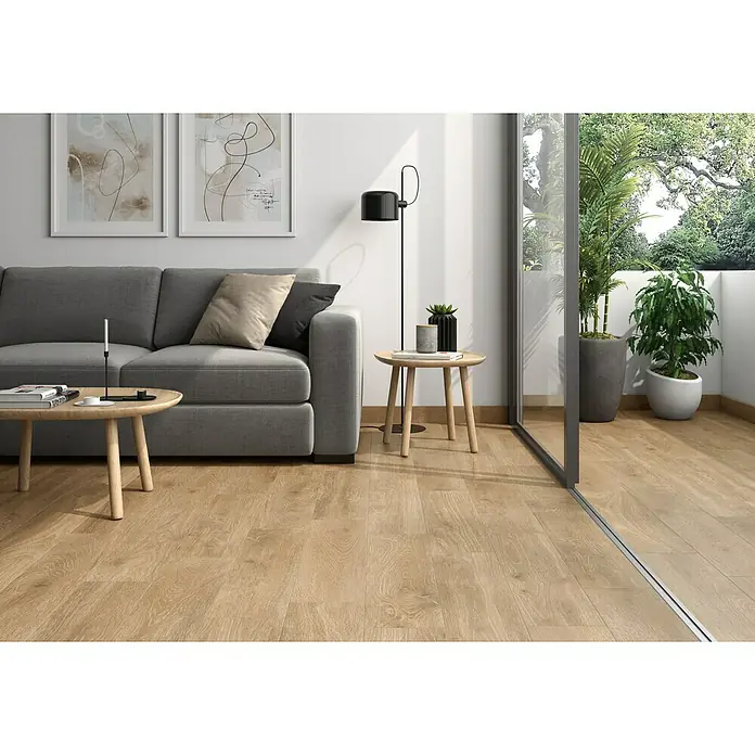 Pavimento porcelánico Galia - 20 x 75 cm, Honey, Mate - Salón con sofá gris, mesa de madera y lámpara de pie. Dos obras de arte enmarcadas adornan la pared sobre el sofá, con plantas en el balcón añadiendo un toque natural.
