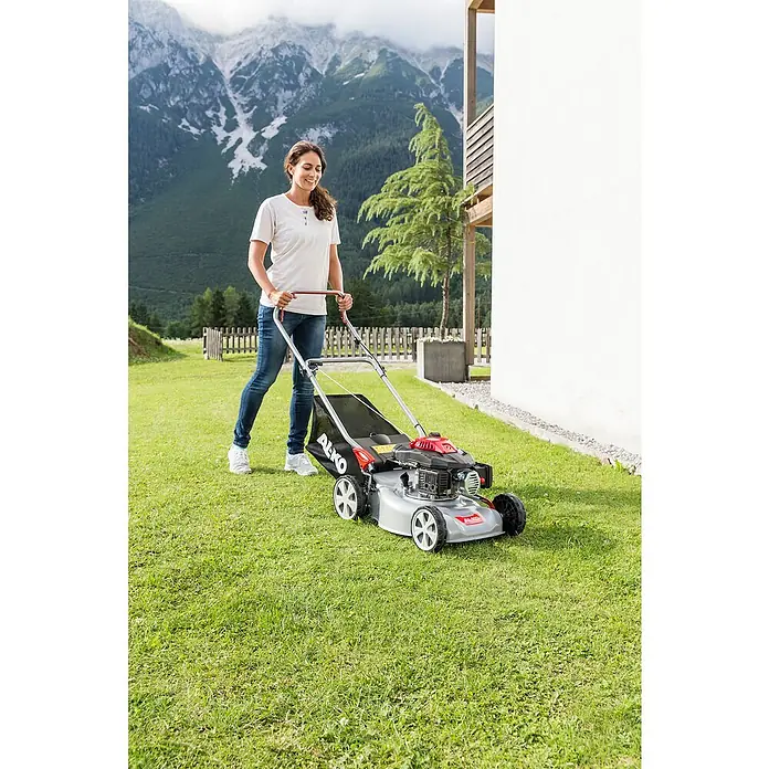 AL-KO Easy Benzin-Rasenmäher 4.20 P-S - 2 kW, 140 cm³, Schnittbreite: 42 cm - Eine Frau mäht den Rasen mit einem AL-KO Rasenmäher vor einer Bergkulisse. Der Rasenmäher ist silberfarben mit roten Akzenten.