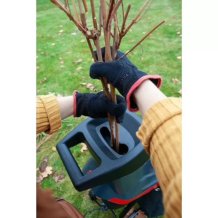 AL-KO Easy Elektro-Häcksler MH2500 Slice - Max. Aststärke: 4 cm, 2.500 W - Person in schwarzen Handschuhen führt braune Äste in einen schwarz-grauen Gartenhäcksler mit roten Akzenten.