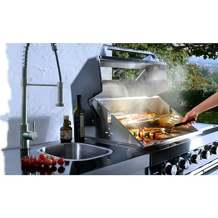 Buschbeck Outdoorküche Cambridge - Schwarz, Material Grillfläche: Edelstahl, Anzahl Brenner: 4 - Edelstahlgrill mit Spüle und Armatur, auf dem Gemüse und Würstchen gegrillt werden. Im Vordergrund liegen reife Tomaten.