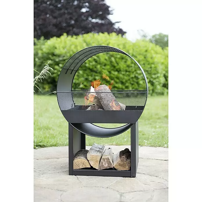 Buschbeck Feuerstelle Porthole - 40 x 56 x 80 cm, Stahl - Schwarze Metall-Feuerstelle mit halbrunder Gitterabdeckung und brennenden Holzscheiten, weitere Scheite darunter gelagert, auf einer Terrasse.