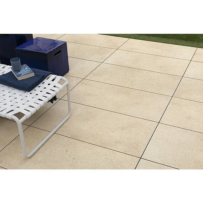 Marazzi Terrassenfliese Esterno 20T Jerusalem Avori50 x 100 x 2 cm, Hellbeige, Matt - Nahaufnahme einer weißen Sonnenliege mit einem Buch und einem blauen Handtuch auf einer gepflasterten Terrasse.