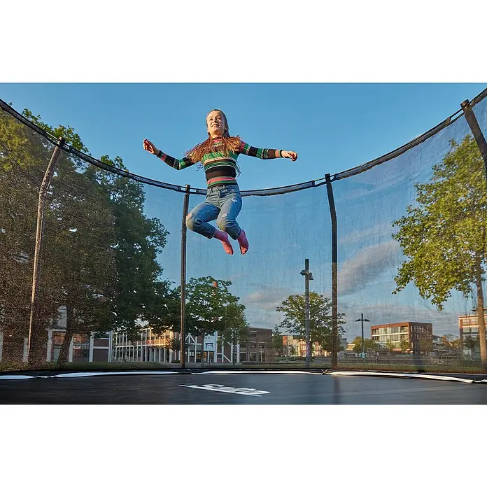 Salta Trampolin Premium Black Edition - Durchmesser: 251 cm, Schwarz - Mädchen springt auf einem Berg Trampolin mit Sicherheitsnetz vor einem Hintergrund von Bäumen und Gebäuden.