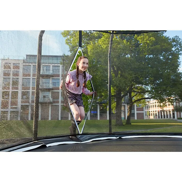 Salta Trampolin Premium Black EditionDurchmesser: 457 cm, Schwarz - Mädchen springt auf einem Trampolin mit Sicherheitsnetz vor einer Baumgruppe und einem Gebäude.