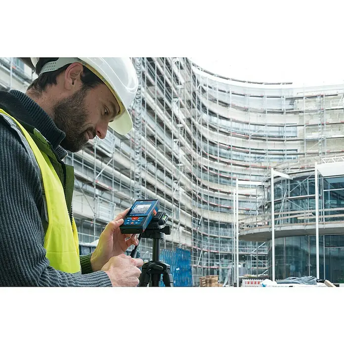 Bosch Professional Laserentfernungsmesser GLM 100-25 C - Messbereich: 0,08 - 100 m - Ein Bauarbeiter mit Schutzhelm bedient einen Bosch-Laser-Entfernungsmesser auf einem Stativ vor einer Baustelle, um genaue Messungen durchzuführen.