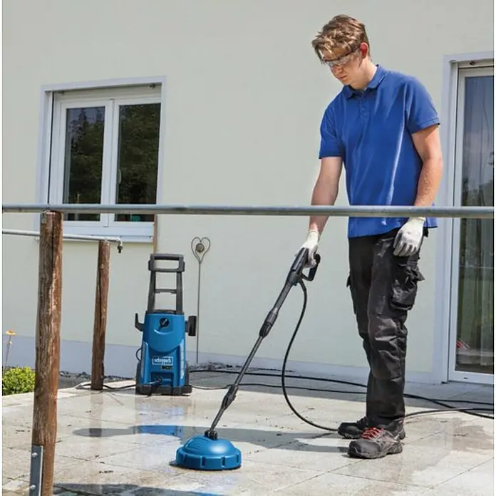 Scheppach Hidrolimpiadora de alta presión HCE 2600 - 180 bar, 2.400 W, Capacidad de bombeo: 480 l/h - Un hombre usa una hidrolimpiadora azul Scheppach con un limpiador de superficies para limpiar un patio.