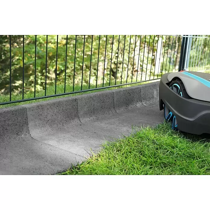 Christoph Mähstein Mähroboter L-Randstein - 24,5 x 29 x 15 cm, Grau, Beton - Grauer und blauer Rasenmähroboter trimmt Gras an einem Betonrand neben einem schwarzen Zaun.