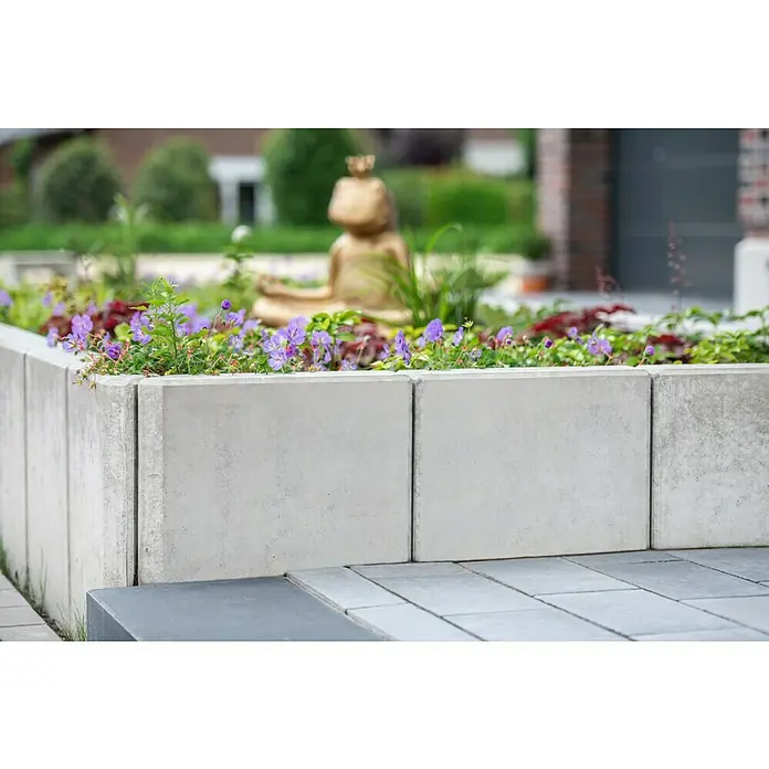Diephaus L-Stein - 55 cm, 100 cm, Stärke: 12 cm, Grau - Nahaufnahme eines erhöhten Blumenbeets aus grauen Betonblöcken, bepflanzt mit lila Blumen und grünen Pflanzen, vor einem Gartenhintergrund.