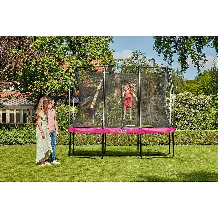 Salta Trampolin Comfort Edition - Pink, L x B: 153 x 214 cm - Ein rechteckiges Gartentrampolin der Marke Salta steht auf einer grünen Wiese, während ein Mädchen darauf springt und zwei Kinder zusehen.