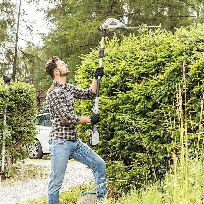 AL-KO 36V Heckenscheren-Aufsatz HTA 4045 - Schnittbreite: 20 cm, Schwertlänge: 45 cm - Ein Mann benutzt eine AL-KO Hochentaster, um eine hohe Hecke zu schneiden. Er trägt Schutzkleidung und arbeitet im Garten.