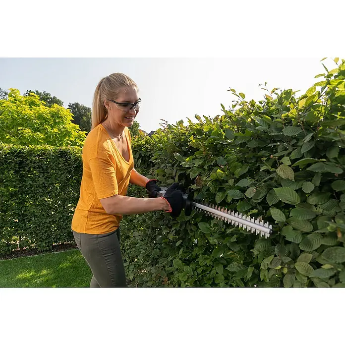 AL-KO Bosch Home & Garden Compatible Akku-Heckenschere HT 1845 - 18 V, Li-Ionen, 1 Akku, Schnittlänge: 45 cm - Eine Frau schneidet eine Hecke mit einer Heckenschere, wobei sie Schutzbrille und Handschuhe trägt, um sich zu schützen.