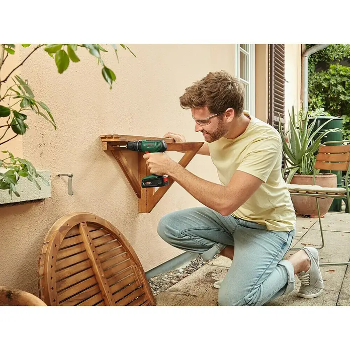Bosch Power for All 18V Maschinenpaket - Akku-Schlagbohrschrauber EasyImpact 18V-40 + gratis Starter-Kit - Mann befestigt eine hölzerne Wandhalterung mit einem Bosch Akku-Bohrschrauber an einer Wand. Er trägt dabei eine Schutzbrille.