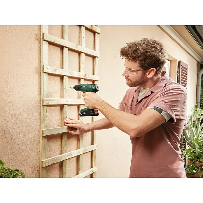 Bosch Power for All 18V Taladro atornillador de batería EasyDrill 18V-40 - 18 V, 1 batería, 2 Ah, 40 Nm - Un hombre sujeta un enrejado de madera a una pared exterior utilizando un taladro inalámbrico Bosch. Lleva gafas protectoras.