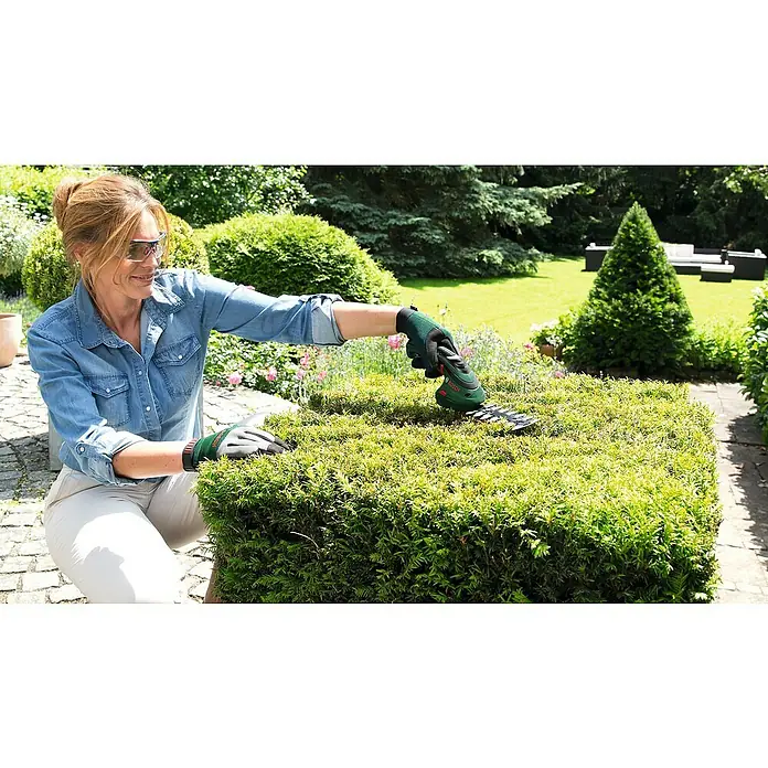 Bosch Podadora de batería - 3,6 V, Iones de litio, 1,5 Ah, 1 batería, Anchura de la hoja: 8 cm, Duración de la batería: 50 min - Una mujer utiliza unas tijeras de arbustos sin cable Bosch para recortar un boj en el jardín, usando guantes y gafas de seguridad.