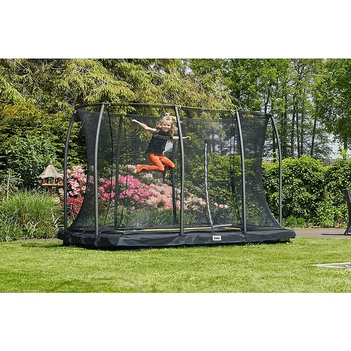 Salta Trampolin Comfort Edition GroundSchwarz, L x B: 153 x 214 cm - Ein Mädchen springt auf einem Salta-Trampolin mit Sicherheitsnetz im Garten, umgeben von üppigem Grün und Blumen, für sicheren Spaß.