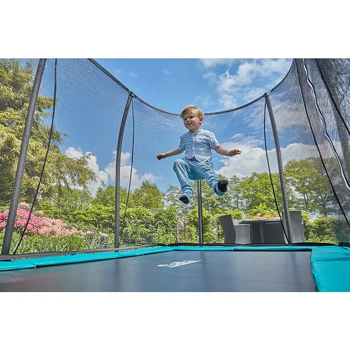 Salta Trampolin Comfort Edition GroundGrün, L x B: 214 x 305 cm - Ein Kind springt auf einem Gartentrampolin mit Sicherheitsnetz vor einem Hintergrund von Bäumen und blauem Himmel.