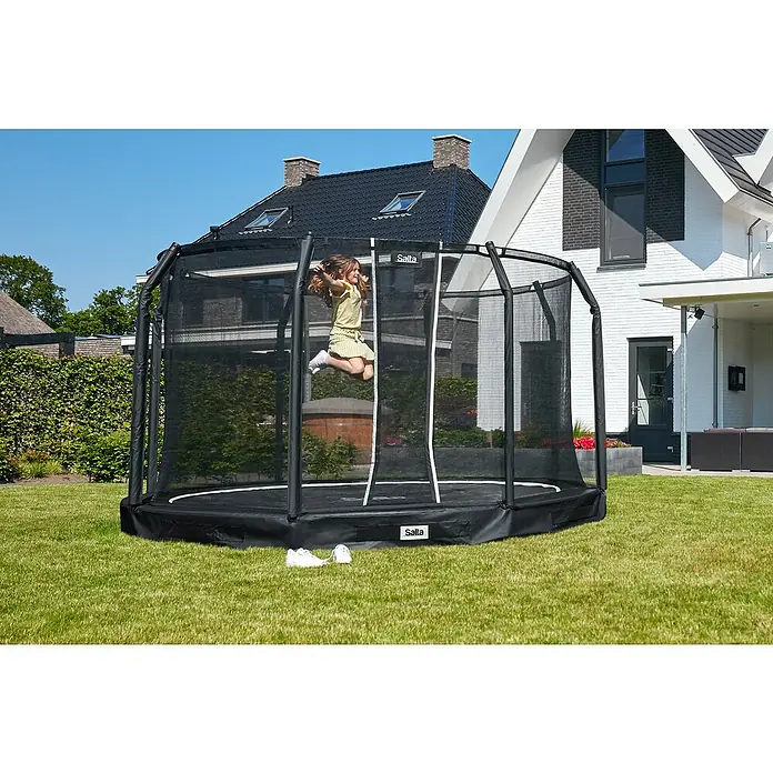 Salta Trampolin Premium Ground - Durchmesser: 251 cm, Schwarz - Ein Salta Trampolin im Garten, auf dem ein Kind springt. Im Vordergrund liegen Schuhe auf dem Rasen. Das Trampolin ist mit einem Sicherheitsnetz ausgestattet.