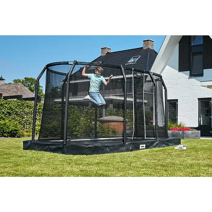 Salta Trampolin Premium Ground - Grün, 214 x 153 cm - Ein Junge springt auf einem rechteckigen Salta-Trampolin mit Sicherheitsnetz im Garten, während seine Schuhe daneben stehen.