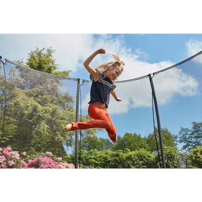 Salta Trampolin Comfort Edition Ground - Schwarz, L x B: 244 x 366 cm - Mädchen springt auf einem Trampolin mit Sicherheitsnetz im Garten, umgeben von üppigem Grün und Blumen.