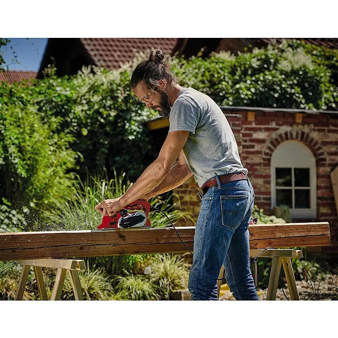 Einhell Expert Elektrohobel TE-PL 900 - 900 W, Hobeltiefe: 0 mm - 3 mm, Leerlaufdrehzahl: 15.000 U/min - Ein Mann hobelt mit einem Einhell Hobel ein Holzbrett im Freien. Das Werkzeug ist rot und schwarz.
