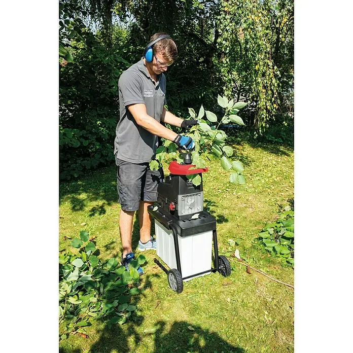 Scheppach Elektro-Leisehäcksler GS60 - Max. Aststärke: 4,5 cm, 2.800 W - Ein Mann benutzt einen Scheppach-Häcksler im Garten, um Äste zu zerkleinern. Er trägt dabei Schutzkleidung für sicheres Arbeiten.