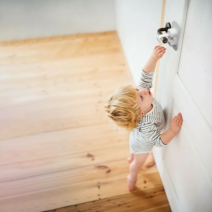 Dreambaby Türsicherung Türklinke - 1 Stk., Weiß - Ein Kleinkind versucht, einen Türgriff mit einem installierten Türgriffschutz zu erreichen, um die Sicherheit zu gewährleisten.