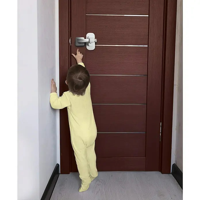 Dreambaby Türsicherung Türklinke - 1 Stk., Weiß - Ein Kind in gelbem Jumpsuit versucht, den installierten Türsicherheitsverschluss zu erreichen, um die Sicherheit zu gewährleisten.