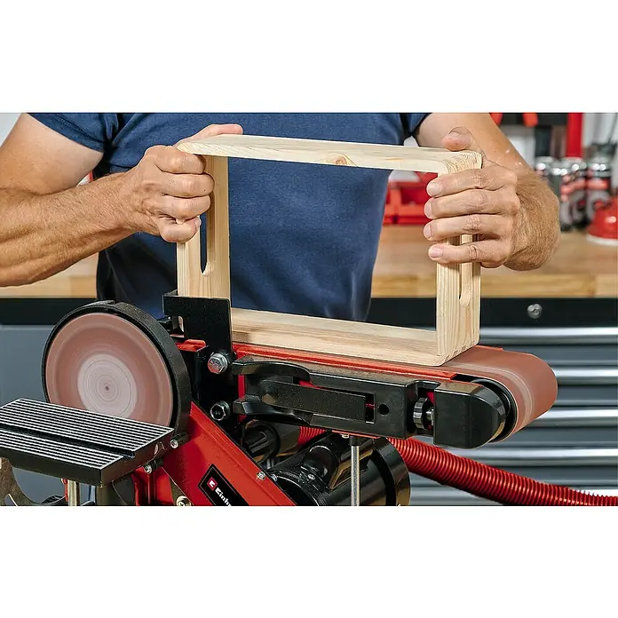 Einhell Schleifband - 914 x 100 mm, Körnung: 60 - 120, 3 Stk. - Eine Person schleift einen Holzrahmen mit einem Bandschleifer. Der Schleifer ist rot und schwarz.