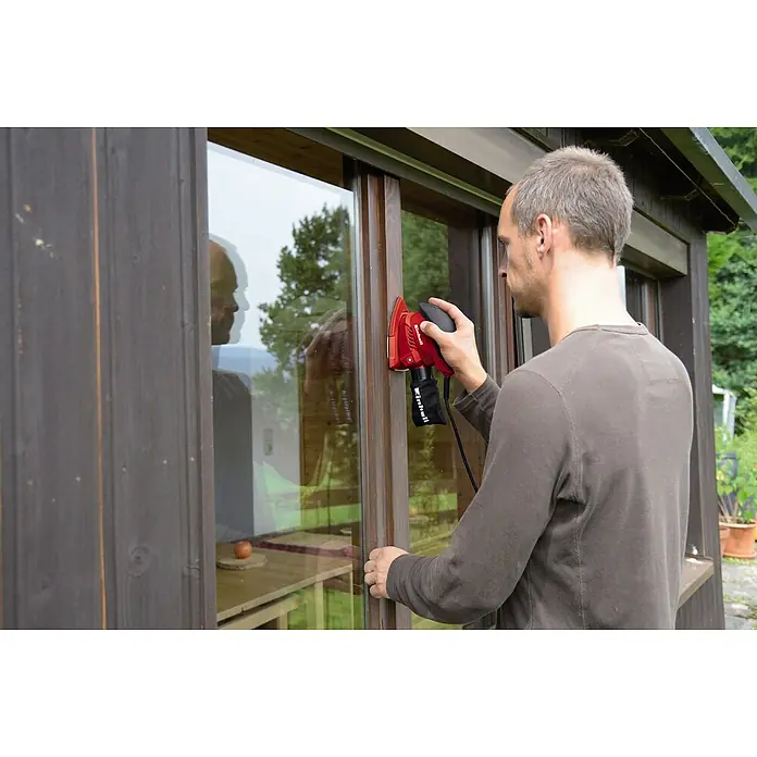 Einhell Elektro-Multischleifer TH-OS 1016 - 100 W, Leerlaufschwingzahl: 24.000 U/min, Durchmesser Schwingkreis: 1,6 mm - Ein Mann schleift mit einem roten Einhell-Schwingschleifer einen braunen Fensterrahmen im Außenbereich.