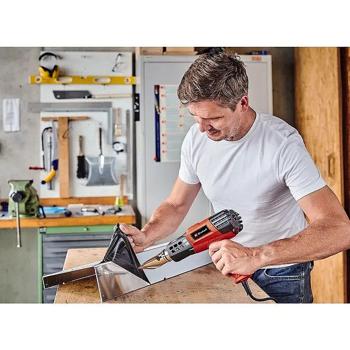 Einhell Expert Heißluftgebläse TE-HA 2000 E - 2.000 W, Temperaturbereich: 50 °C - 550 °C - Ein Mann verwendet eine Einhell Heißluftpistole, um eine Ecke eines Metallgegenstandes zu bearbeiten. Die Pistole ist rot und schwarz.