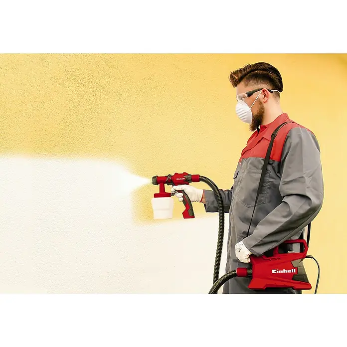 Einhell Classic Farbsprühsystem TC-SY 600 S - 600 W, Förderleistung: 1 l/min - Ein Mann sprüht mit einer roten Einhell-Farbspritzpistole eine gelbe Wand weiß. Die Spritzpistole ist mit einem Schlauch mit einer Basis verbunden.
