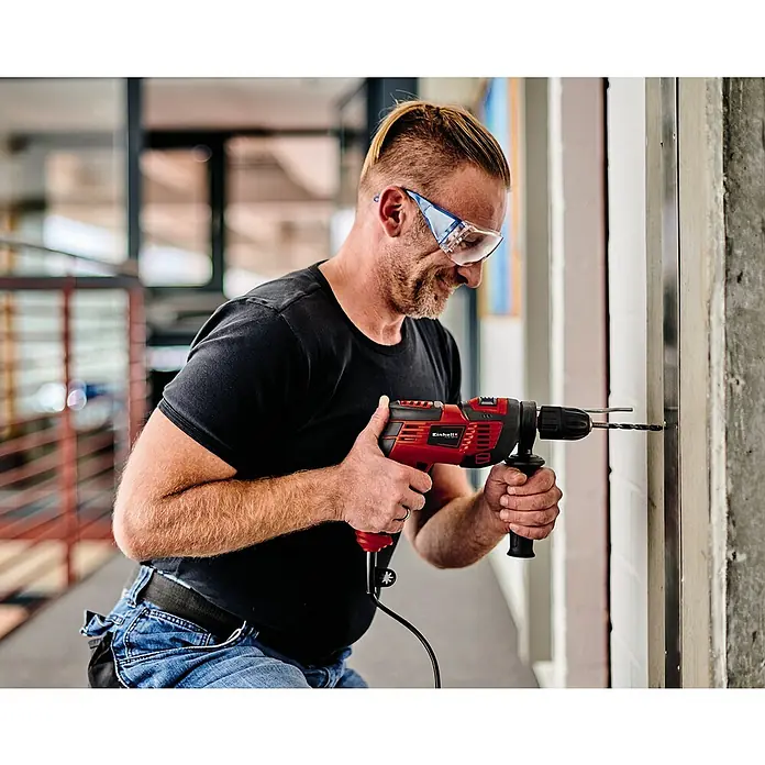 Einhell Schlagbohrmaschine TC-ID 720/1 E - 720 W, 2.800 U/min, Max. Schlagzahl: 44.800 /min - Ein Mann bohrt mit einer roten Einhell Schlagbohrmaschine ein Loch in eine Wand, Schutzbrille inklusive.