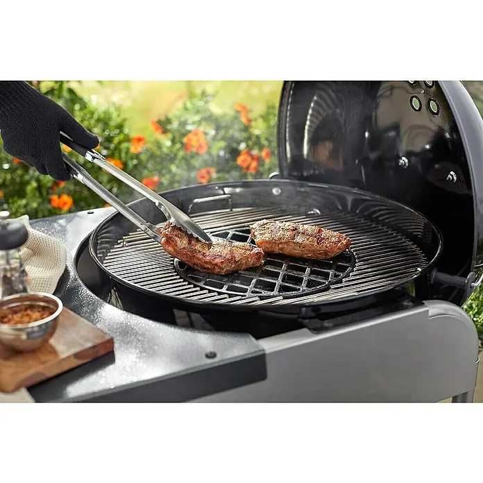 Weber Gourmet BBQ System Grillrost Sear Grate - L x B: 30 x 30 cm, Durchmesser: 30 cm, Gusseisen - Ein Grill mit zwei Steaks darauf, die mit einer Zange gewendet werden. Im Hintergrund sind Blumen zu sehen.