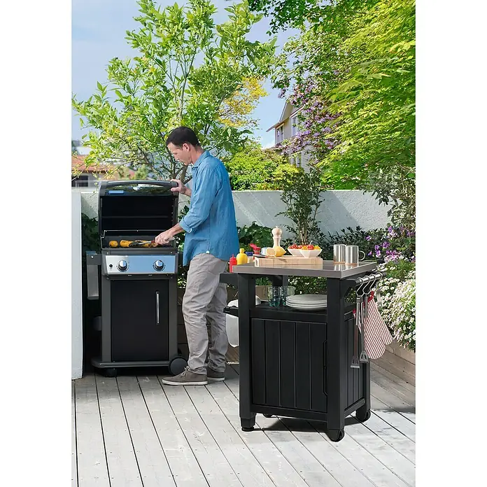 Keter Mesa auxiliar de jardín Unity - L x An: 82 x 52 cm, Marrón - Un hombre asando maíz en una parrilla de gas al aire libre, con una mesa auxiliar con ingredientes y accesorios cerca.