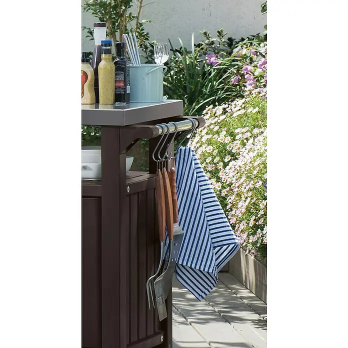 Keter Mesa de servicio Unity XL - 123,7 x 54 cm, Plástico, Marrón - Primer plano de un carrito de parrilla con utensilios para asar y una toalla de rayas azules y blancas.