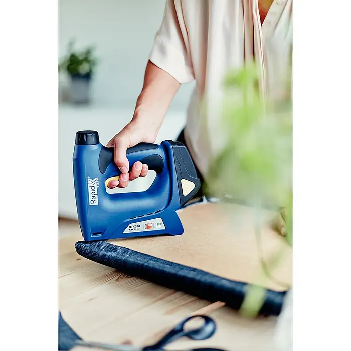 Rapid Akku-Tacker BTX530 - 7,2 V, Li-Ionen, 1,3 Ah, 1 Akku, Klammertiefe: 6 mm - 14 mm - Nahaufnahme einer Person, die einen blauen Rapid Btx530 Akku-Tacker auf einem Holztisch bedient.