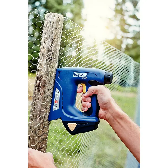 Rapid Akku-Tacker BTX530 - 7,2 V, Li-Ionen, 1,3 Ah, 1 Akku, Klammertiefe: 6 mm - 14 mm - Ein Rapid BXT530 Akku-Tacker befestigt Drahtgeflecht an einem Holzpfosten. Das blaue Werkzeug liegt gut in der Hand und sorgt für sicheren Halt.