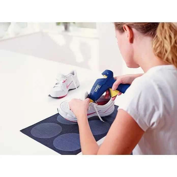 Rapid Set para pistola termoadhesiva EG212 - 200 W, Rango de temperaturas: 170 °C - 190 °C - Mujer usando una pistola de pegamento Rapid para reparar una zapatilla deportiva blanca con detalles rosas, otro zapato al fondo.